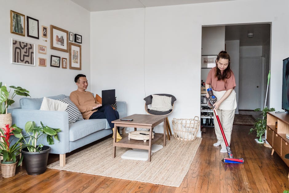 A woman with brown hair tied back, wearing a pink blouse and beige trousers, is operating a cordless vacuum cleaner with a red wand in a residential living area. She is standing on a polished wooden f