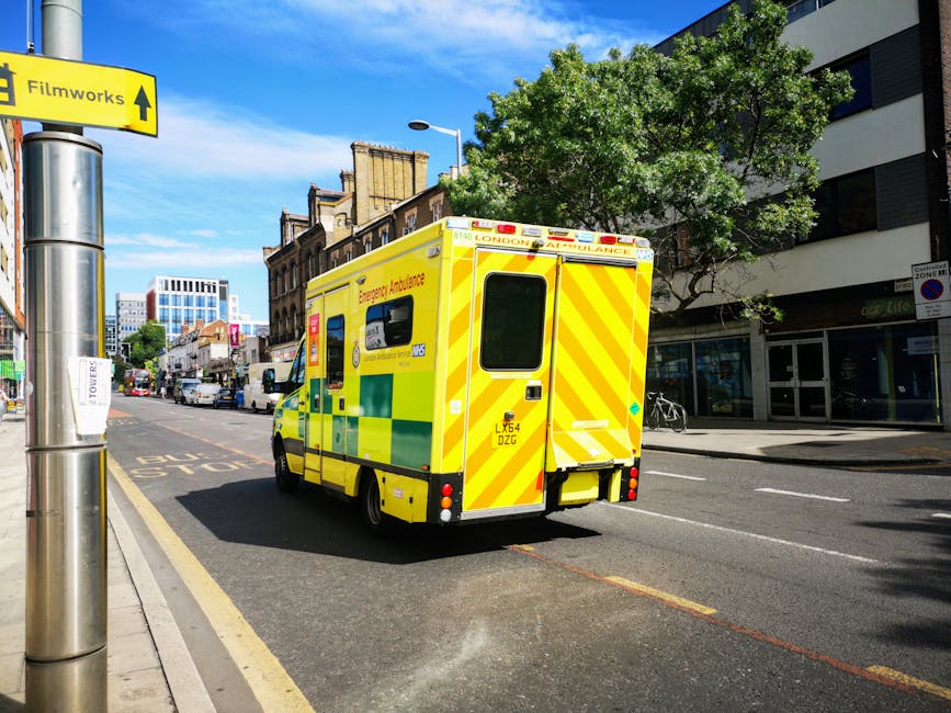 A bright yellow emergency ambulance with green and reflective safety markings is parked on the street near a modern building with large windows and a white facade. The ambulance features a rear door with a window, and the vehicle's lights are off. The street scene includes a tree with lush green foliage casting shadows on the pavement, a bicycle locked to a railing, and other vehicles in the distance. On a lamppost in the foreground, a yellow sign with black text points towards the Filmworks area. The sky is clear and blue, indicating a sunny day. This image captures a typical urban scene in Maida Vale, with the emergency vehicle ready for rapid response, and the surroundings are clean and well-maintained, exemplifying the borough's infrastructure. Cleaners Maida Vale specialises in thorough cleaning services, ensuring hygiene and cleanliness in both residential and commercial environments, reflected in the bright, orderly street scene.
