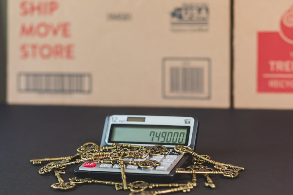 A close-up view of a calculator displaying the number 749800, surrounded by several gold-colored keys and keychains, resting on a dark surface. In the background, there are blurred cardboard boxes with red and blue printed labels indicating shipping or storage instructions. The scene is evenly lit, emphasizing the metallic shine of the keys and the digital screen of the calculator. This image visually supports the concept of cost assessment or valuation related to professional cleaning services provided by Cleaners Maida Vale for end of tenancy cleaning in Maida Vale W9, focusing on surface cleaning and cost estimation for domestic cleaning projects.