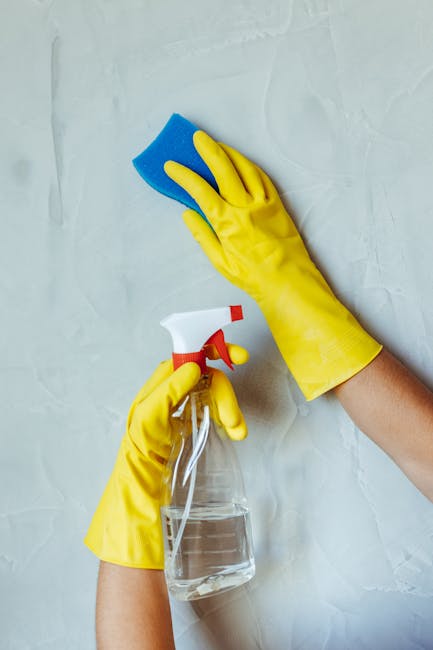 Close-up of a cleaning scene showing hands wearing bright yellow rubber gloves holding a blue kitchen sponge, poised to clean a glass spray bottle with a red and white spray top, placed on a light gra
